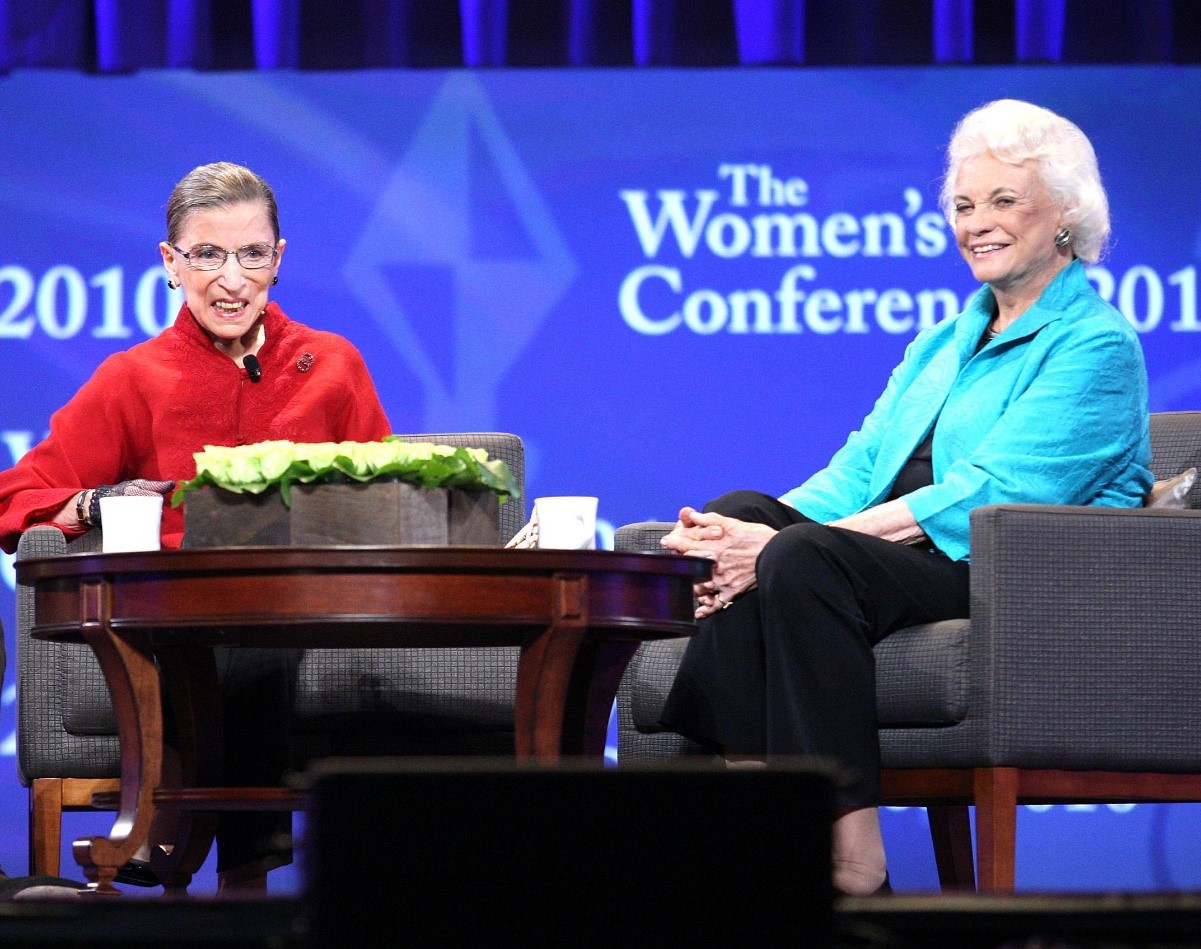 Two women sit on stage at a conference, engaging in conversation. One wears a red top and glasses, the other wears a turquoise jacket. They're seated at a small table with a plant, against a backdrop displaying "The Women's Conference.