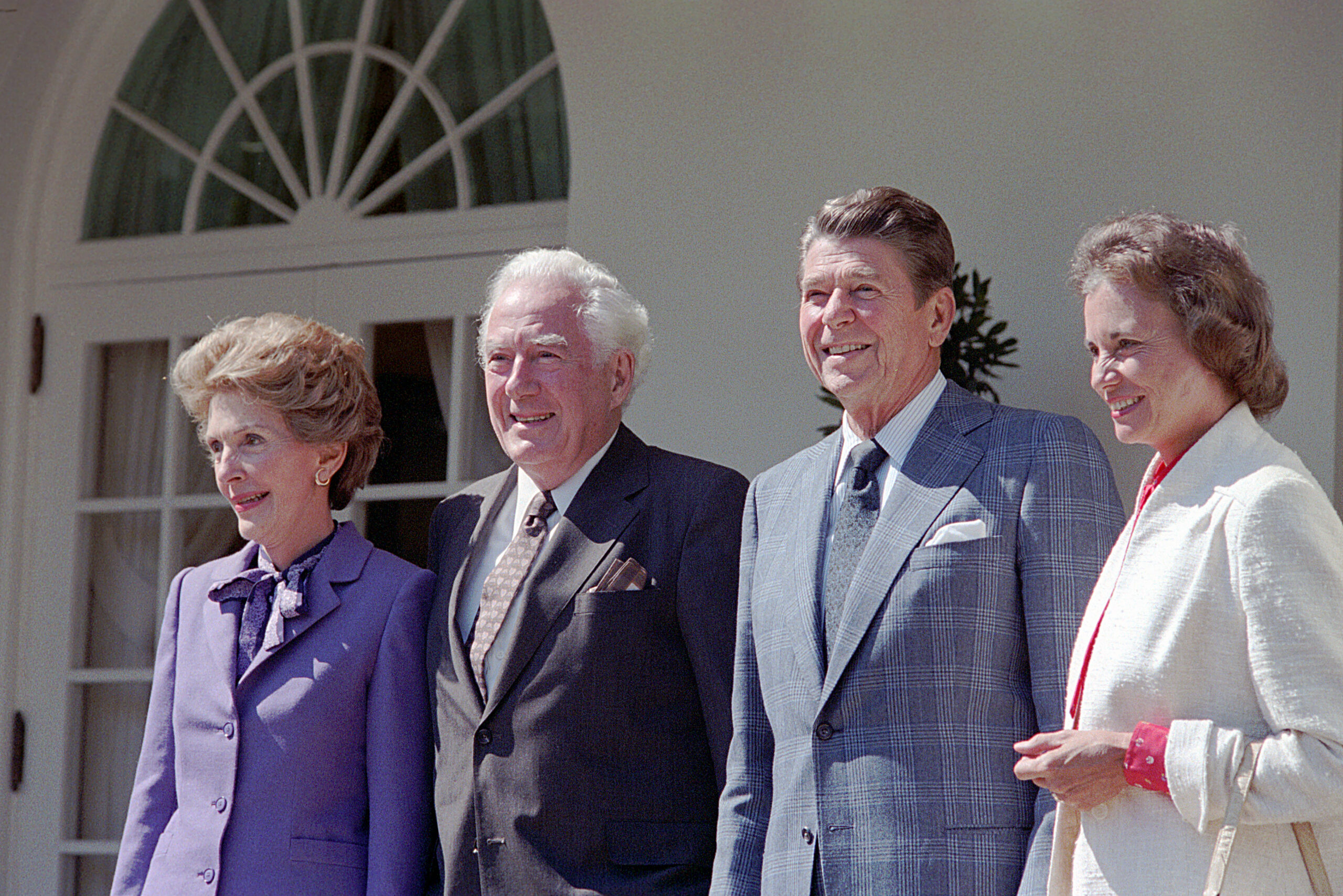 Four adults, two men and two women, stand side by side outside a white building with an arched window, smiling for the camera. The men wear suits and ties; the women wear formal jackets.